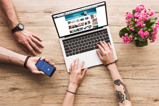 cropped shot of man with smartphone with facebook logo in hand and woman at tabletop with laptop with amazon website and kalanchoe flower - Zdjęcie, obraz