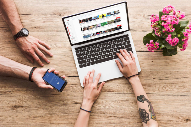 cropped shot of man with smartphone with facebook logo in hand and woman at tabletop with laptop with youtube website and kalanchoe flower - Fotó, kép