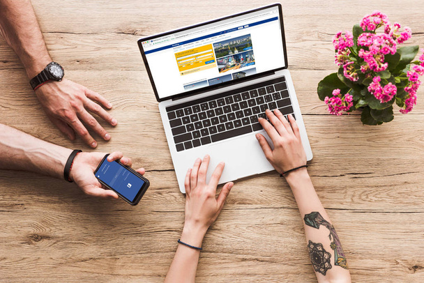 cropped shot of man with smartphone with facebook logo in hand and woman at tabletop with laptop with bookingcom website and kalanchoe flower - Photo, Image