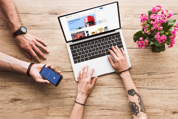 cropped shot of man with smartphone with facebook logo in hand and woman at tabletop with laptop with ebay website and kalanchoe flower - Valokuva, kuva