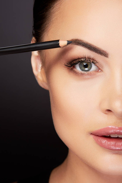 Schönheitskonzept, junge Frau schminkt sich, streicht Augenbrauen mit schwarzem Bleistift, schwarzer Hintergrund - Foto, Bild