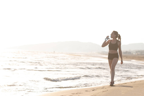 Gezonde jonge passen vrouw drinkwater wandelen op zonsopgang aan zee trail - Foto, afbeelding