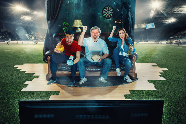 Soccer football fans sitting on the sofa and watching TV in the middle of a football field. - Zdjęcie, obraz