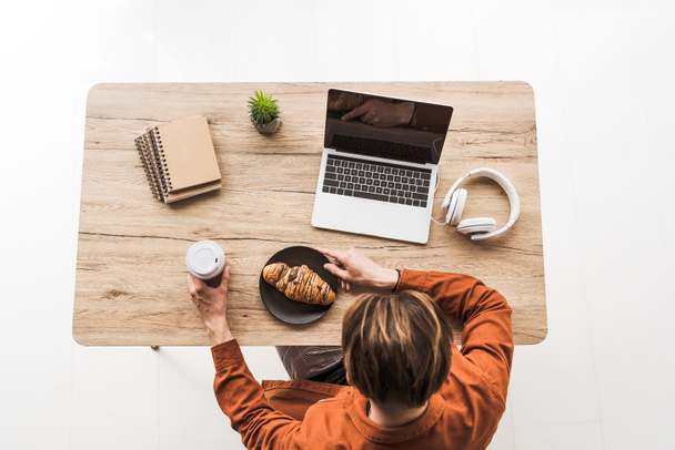 vue surélevée de l'homme avec café et croissant travaillant à table avec ordinateur portable, écouteurs, manuels scolaires et plantes en pot
 - Photo, image