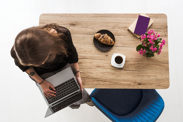 overhead view of woman typing on laptop and sitting on table with croissant, coffee, flowers, book and textbook  - Fotó, kép