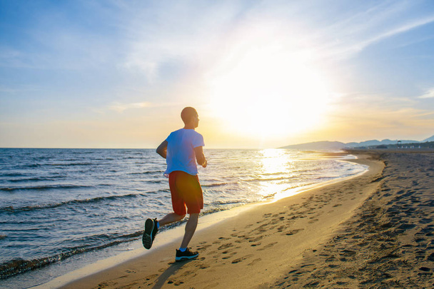 Rückansicht eines gesunden Mannes, der am Strand gegen den Sonnenuntergang joggt - Foto, Bild