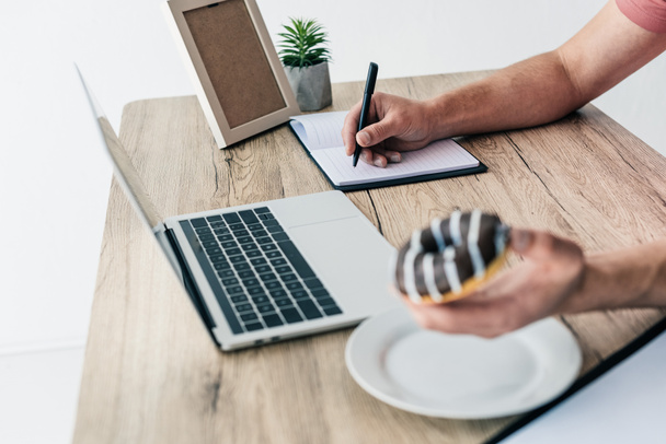 περικοπεί shot του ανθρώπου με ντόνατ γράφοντας στο βιβλίο στο τραπέζι με το laptop, κορνίζα και γλάστρες φυτών  - Φωτογραφία, εικόνα