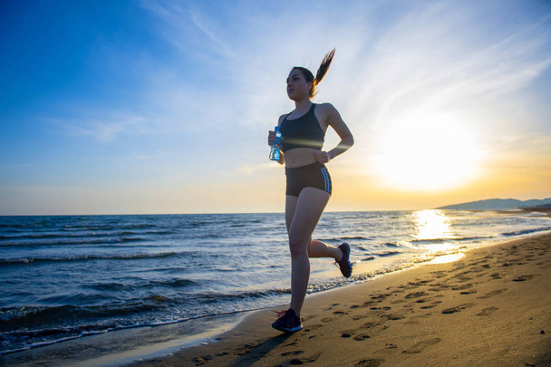 Vista lateral de longitud completa de la mujer en forma sosteniendo botella de agua corriendo en la playa al atardecer
 - Foto, Imagen