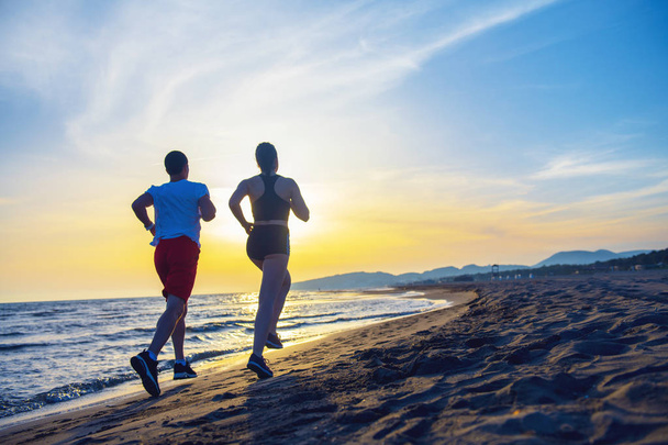 Visão traseira de comprimento total do casal correndo juntos na praia contra o céu do pôr do sol
 - Foto, Imagem