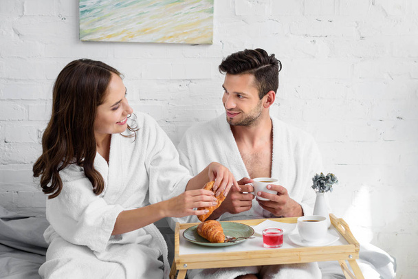 young couple in bathrobes having croissants and coffee for breakfast in bed - Fotó, kép