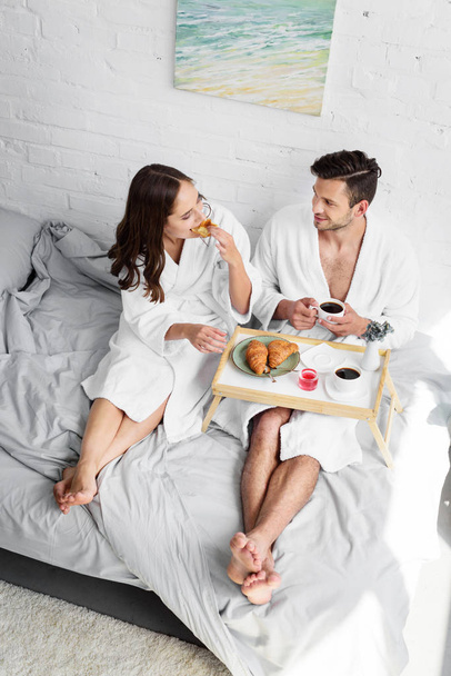 beautiful girlfriend and boyfriend in bathrobes having croissants and coffee for breakfast in bedroom - Fotografie, Obrázek