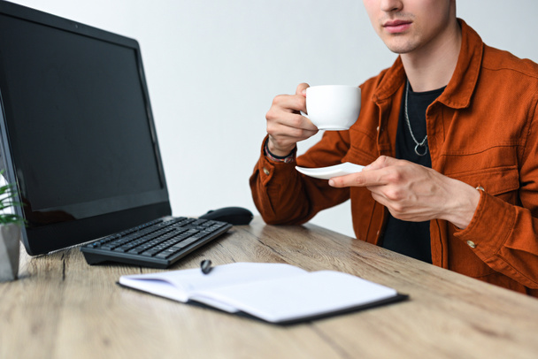 vista parcial del hombre sosteniendo la taza de café en la mesa con el libro de texto, computadora, teclado de la computadora y ratón de la computadora
 - Foto, imagen