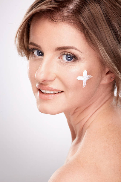 pretty smiling young woman with naked shoulders using facial cream, beauty photo concept, skin and wrinkles treatment, UV protection, plus symbol on skin  - Photo, Image