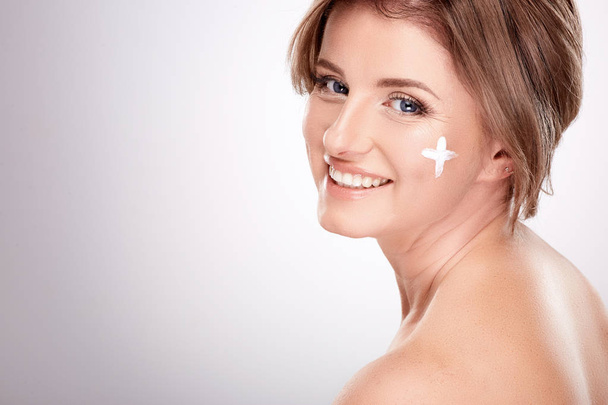 hübsch lächelnde junge Frau mit nackten Schultern mit Gesichtscreme, Schönheitsfotokonzept, Haut- und Falten-Behandlung, UV-Schutz, plus Symbol auf der Haut  - Foto, Bild