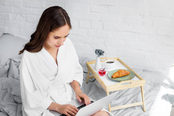 attractive young woman in white bathrobe using laptop during breakfast in bedroom - Foto, afbeelding