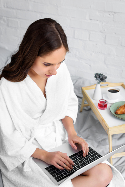 krásná žena v bílém županu pomocí přenosného počítače během snídaně v posteli - Fotografie, Obrázek