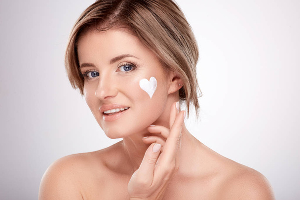 pretty smiling young woman with naked shoulders using facial cream, beauty photo concept, skin and wrinkles treatment, UV protection, heart symbol on skin  - Photo, Image