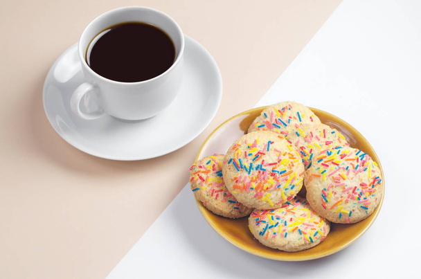 Kuppi kahvia ja maukkaita evästeitä värikkäillä sprinkleillä levyllä valkoisella ja vaaleanruskealla taustalla
 - Valokuva, kuva