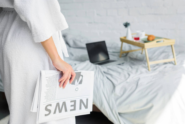 vista parcial de la mujer con el periódico de pie cerca de la cama con desayuno y portátil
 - Foto, Imagen