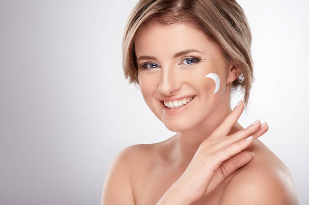 pretty smiling young woman with naked shoulders using facial cream, beauty photo concept, skin and wrinkles treatment, UV protection, crescent symbol on skin  - Фото, зображення