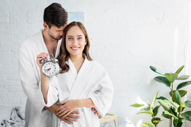 couple in white bathrobes embracing and holding alarm clock in morning - Фото, изображение