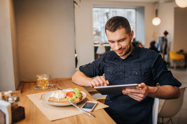 A picture of businessman sitting at table and working. He is using tablet. There is a plate with tasty food and drink on the table. Also there is a phone lying near plate. - Foto, imagen