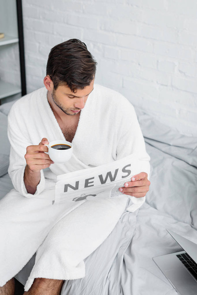 young man in bathrobe sitting on bed, reading newspaper and holding cup of coffee - Foto, immagini