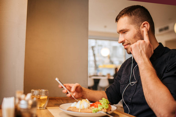 Nice and attractive guy is sitting at tabel and holding phone in his hands. He is listening to music via headphones. Guy is holding forefinger close to ear and looking at phone. - Foto, afbeelding