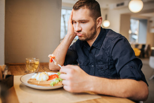 Bored and sad man is sitting at table and cafe. He is holding a piece of vegetable on fork. Man is looking at it and breathing out. - Foto, Imagem
