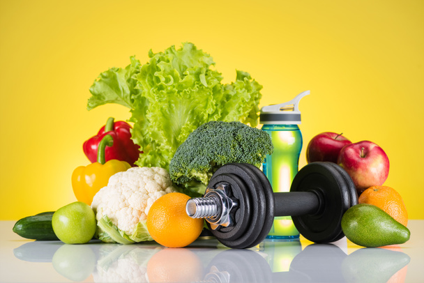 close-up view of dumbbell, bottle of water and fresh fruits and vegetables on yellow  - Photo, Image