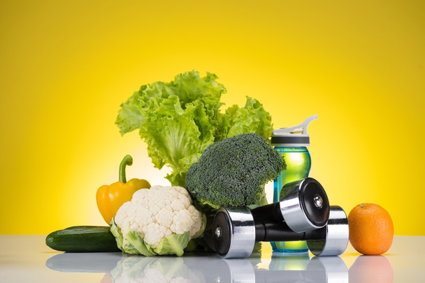 verse gezonde groenten, fles water op geel, oranje en halters - Foto, afbeelding