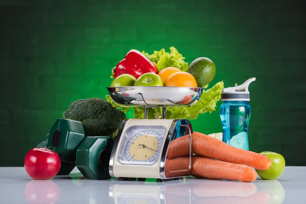 close-up view of fresh fruits and vegetables on scales, dumbbells and bottle of water on green - Photo, Image
