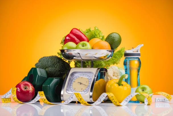 close-up view of fresh fruits and vegetables on scales, sports bottle with water, dumbbells and measuring tape on yellow   - Photo, Image