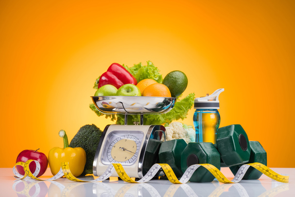 fresh fruits and vegetables on scales, sports bottle with water, dumbbells and measuring tape on yellow   - Photo, Image