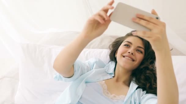Taking selfie chatting with mobile phone beautiful young smiling woman in bed. Video footage - Filmagem, Vídeo