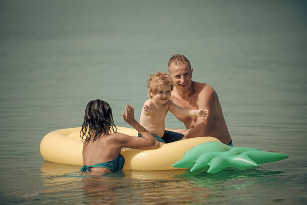 Father and mother near mattress swim with son. Family spend time together and having fun. Cute child boy sits on air mattress pineapple shaped in the ocean, sea, with parents. Family vacation concept - Φωτογραφία, εικόνα