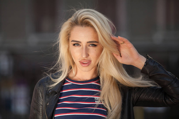 Close-up portrait of young blonde girl with beautiful eyes wearing black jacket outdoors. Pretty female with long wavy hair hairstyle. Woman in urban background. - Fotó, kép