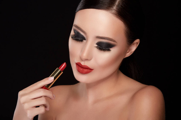 portrait of beautiful young woman with dramatic make up painting lips with red lipstick against black studio background, fake eyelashes - Фото, зображення