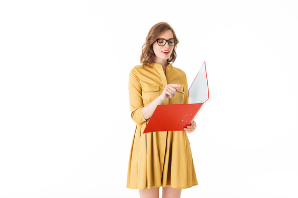 Portrait of pretty thoughtful lady in eyeglasses and yellow dress standing with red folder in hand on white background isolated - Photo, Image