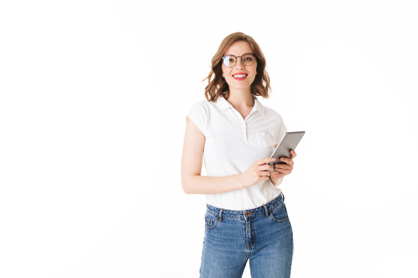 Portrait of young beautiful lady in eyeglasses standing with the tablet in hands and happily looking in camera on white background isolated - Photo, Image