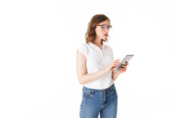 Portrait of beautiful lady in eyeglasses standing and thoughtfully working on the tablet on white background isolated - 写真・画像