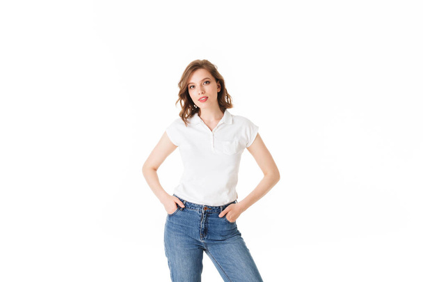 Портрет молодой красивой девушки, стоящей в футболке и джинсах и мечтательно смотрящей в камеру на белом фоне изолированной
 - Фото, изображение