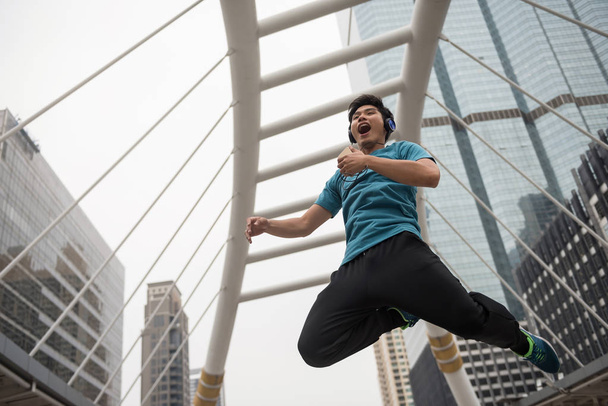 Happy athlete young man with sport cloth jump while listen music by smartphone with Modern Bangkok building background. Exercising outside with full energy. Happy health lifestyle concept - Photo, Image