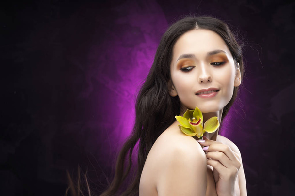 A beautiful brunette nude shoulders girl sensually holds a flower of an orchid in her hands. Nude make-up. Healthy smooth skin. Purple-black background. Copy space. Commercial and advertising. - Photo, image