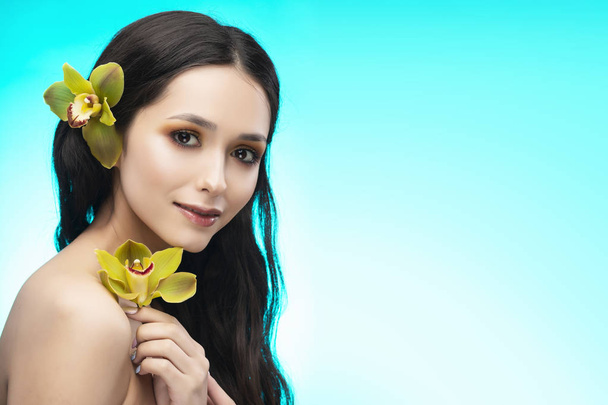 A beautiful brunette nude shoulders girl sensually holds a flower of an orchid in her hands. Nude make-up. Healthy smooth skin. Sky blue background. Copy space. Commercial and advertising design. - Photo, Image