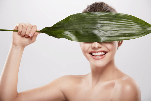 schöne und natürliche junge Frau posiert mit großem grünen Blatt vor grauem Hintergrund, Hautpflegekonzept, hydratisierte Haut - Foto, Bild