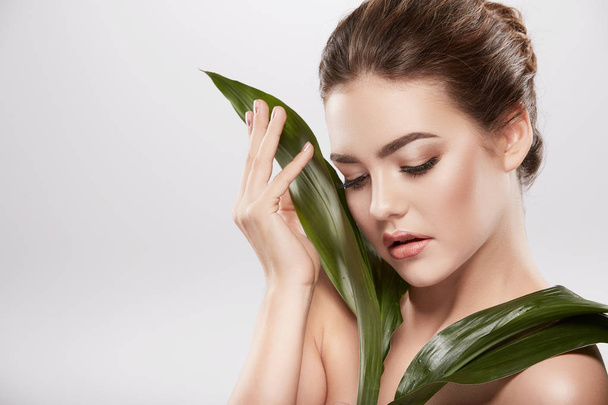 beautiful and natural young woman posing with green leaves at grey background, skin care concept, hydrated skin - Photo, Image