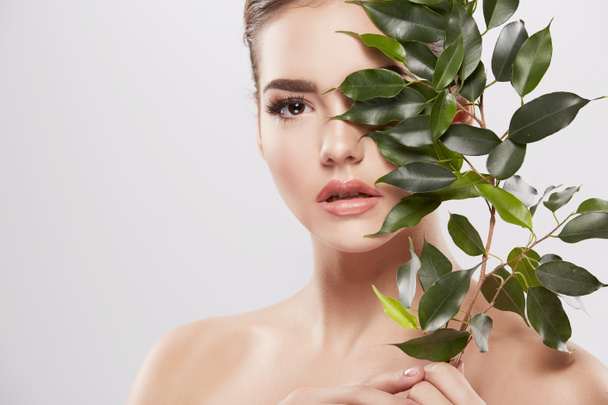 mooie en natuurlijke jonge vrouw poseren met groene bladeren op een grijze achtergrond, huid zorg concept, gehydrateerde huid - Foto, afbeelding