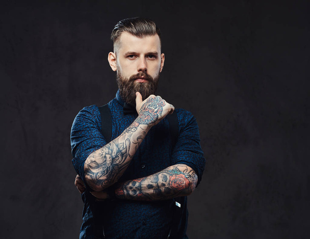 Συλλογισμένος όμορφος ντεμοντέ hipster σε ένα μπλε πουκάμισο και ζαρτιέρες, στέκεται με το χέρι στο πηγούνι σε ένα στούντιο. - Φωτογραφία, εικόνα