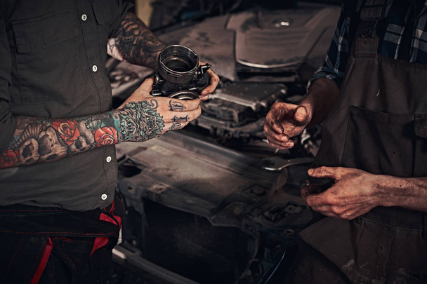 Zwei Mechaniker untersuchen ein Detail des Motors, während sie neben einem kaputten Auto in einer Garage stehen. - Foto, Bild